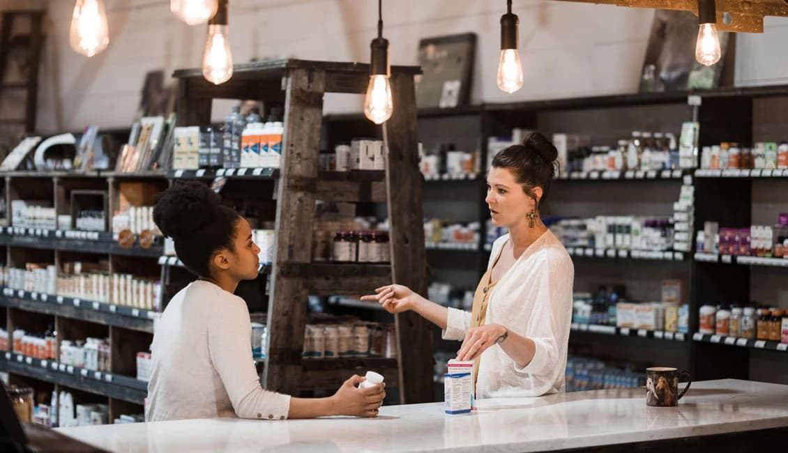A staff member talking a customer through a supplement at the counter, shelves of vitamins behind them