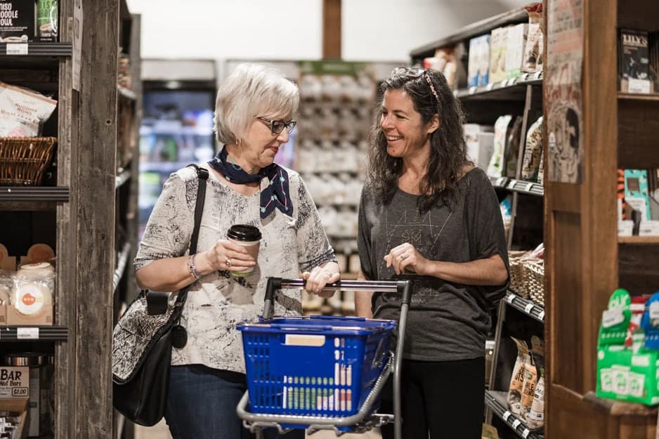 Two shoppers chatting in the aisle, one holding a coffee and a basket of groceries